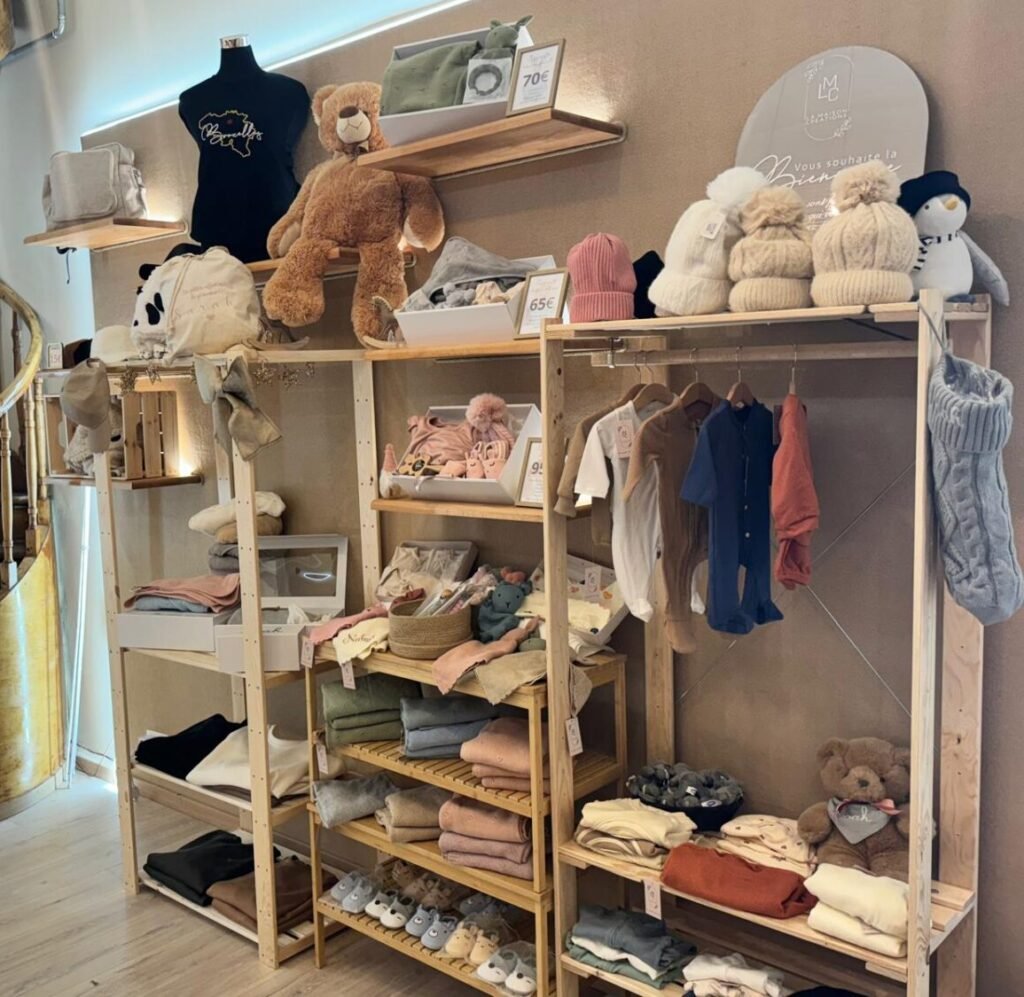 Option 2 (Très Descriptive) Ambiance chaleureuse dans une boutique pour enfants avec des étagères et des portants en bois clair. On y voit une sélection organisée de vêtements pour bébés, de bonnets en tricot, de chaussons, de couvertures pliées et de peluches. Un panneau décoratif indique "La Maison Créations" et une ambiance accueillante, illustrant une offre diversifiée de cadeaux de naissance personnalisés.