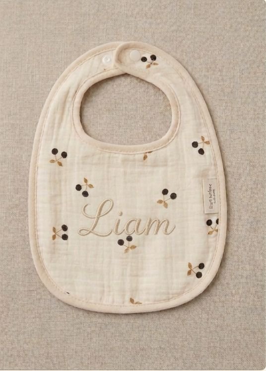 Bavoir pour bébé en tissu mousseline beige clair, parsemé d'un motif de petites cerises, avec une personnalisation brodée au prénom 'Liam' au centre et une étiquette de marque sur le côté.