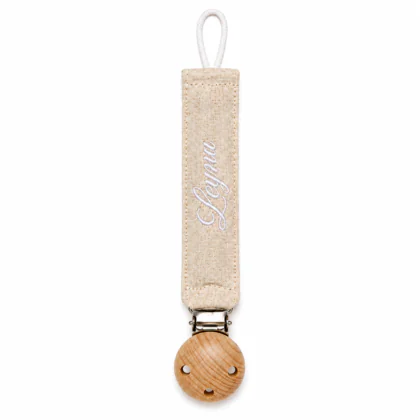 Attache-tétine pour bébé composé d'une sangle plate en tissu beige clair brodé du prénom "Leyna" en blanc, d'une attache métallique et d'un embout rond en bois naturel avec trois petits trous décoratifs.