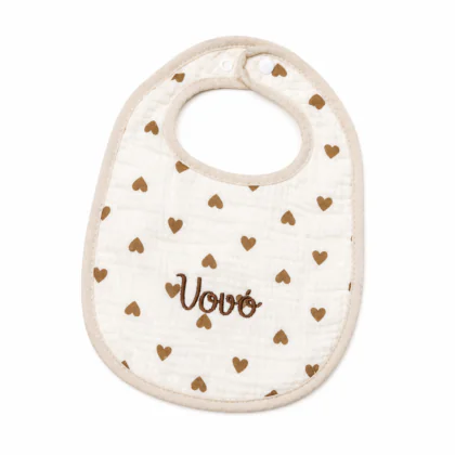 Bavoir pour bébé en tissu mousseline blanc cassé, orné d'un motif répétitif de petits cœurs marron. Le mot "Vovó" est brodé en lettres cursives marron foncé au centre, avec un liseré beige et une fermeture par bouton pression.